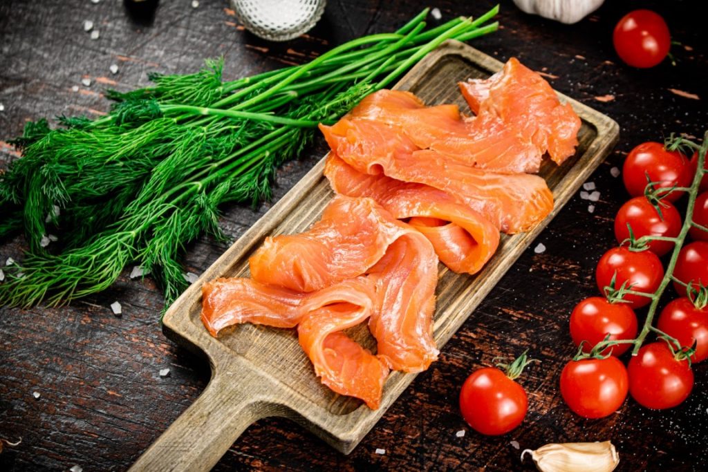 Slices of salted salmon on a cutting board with dill and tomatoes.
